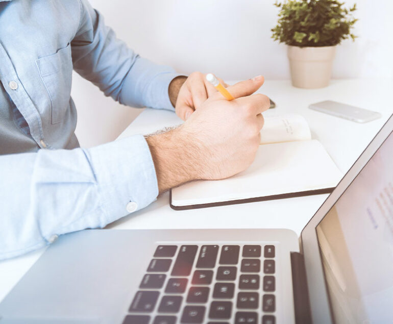 Person holding a pencil, writing in a notebook beside a laptop and plant.
