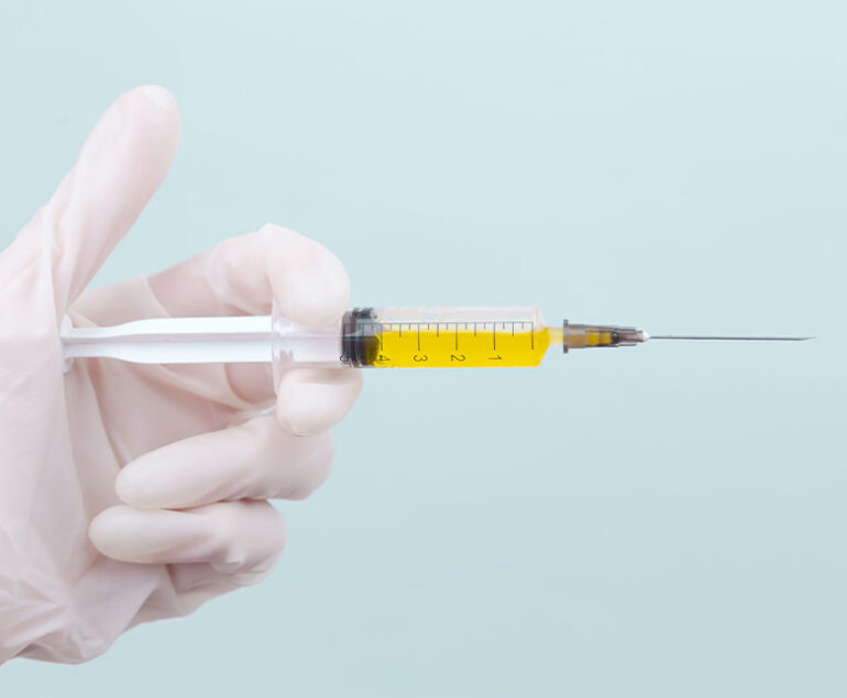 Gloved hand holding a syringe filled with yellow liquid against a light blue background.